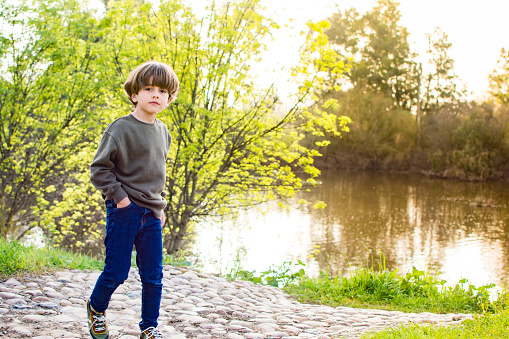 Little Boy Walking In The Field Next To A River At Sunset. Nature And Lifestyle