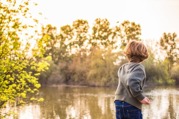 Child Throwing Stones Into The River. Boy Playing By The River At Sunset. Lifestyle And Nature
