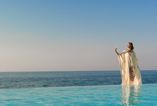 Greek style woman with glass of wine stand on the edge of infinity pool near seaside