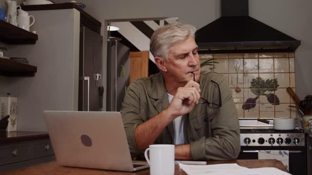 Caucasian mature male working in kitchen on laptop drinking coffee