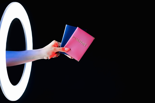 Passport control. Visa service. Foreign embassy. Female hand holding two citizenship ID in pink blue cover in LED light circle portal isolated on black empty space background.