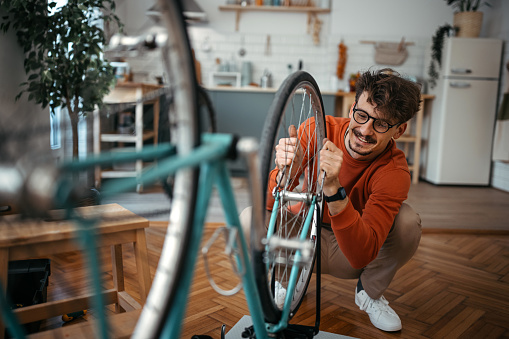 Handsome young man wearing orange sweatshirt adjusting bicycle tire while crouching at home