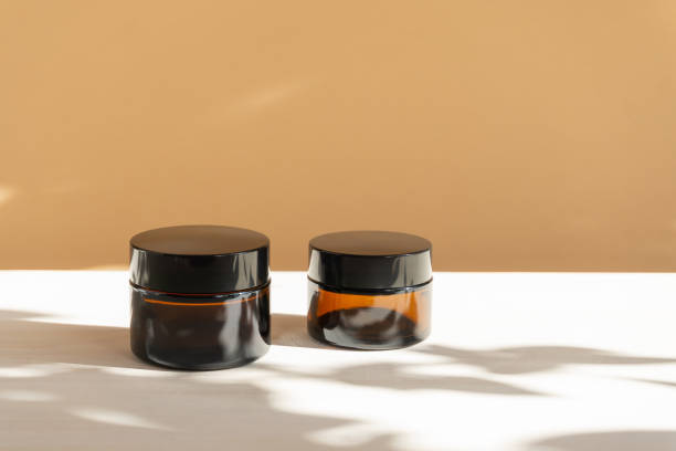 istock Mock-up of amber-colored glass jars for moisturizer on beige background in sunlight. Container with cosmetic products for face in close-up and with floral shadow 1382201325
