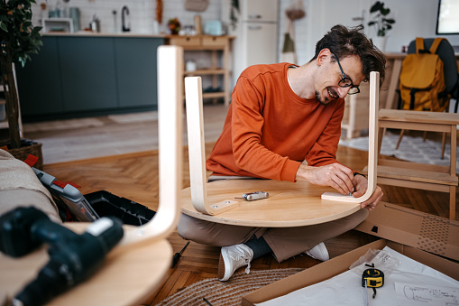 Handsome young man wearing eyeglasses installing wooden table with drill while sitting at home