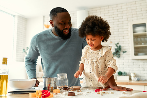 African American family with their little daughter with curly fluffy hair having fun and cooking pastries in the kitchen. Dad and daughter cooking together and cutting out cookies from the dough.