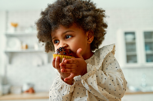 An African American little baby with curly fluffy hair in a dress eats a chocolate muffin with appetite in the kitchen at home.