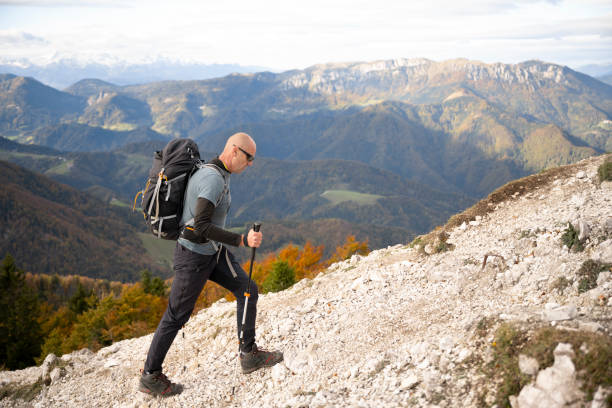 Side view of mature man hiking with hiking poles on dirt track on mountain against cloudy sky.