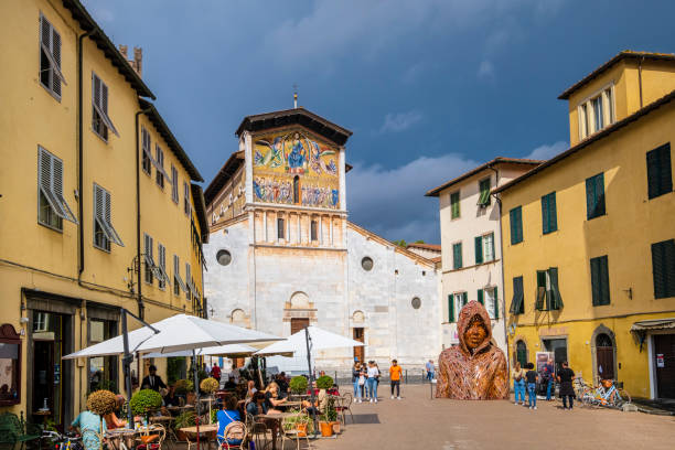 People sitting at the outdoor tables in front of the Basilica of San Frediano, a Romanesque church completed in the 12th century with a monumental golden mosaic on the façade. In the square stands out the sculpture titled 'Hoodie', an artwork of Michael Stutz installed at the 2016 edition of the Lucca Biennale.