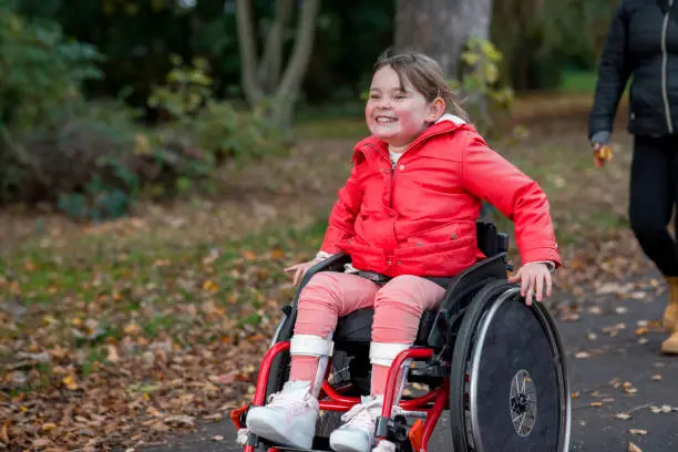 A low-angle view of a young girl out with her family for a day out in a public park in Newcastle upon Tyne. She is a wheelchair user and is pushing herself ahead of her family in the park.