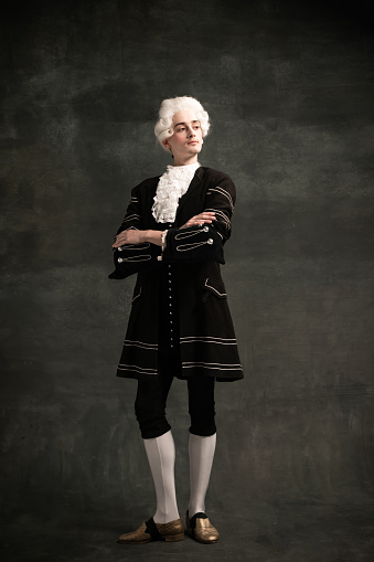 Aristocrat. Young man wearing wig and vintage medieval outfit like famous composer isolated on dark green vintage background. Retro style, fashion, art, comparison of eras concept.