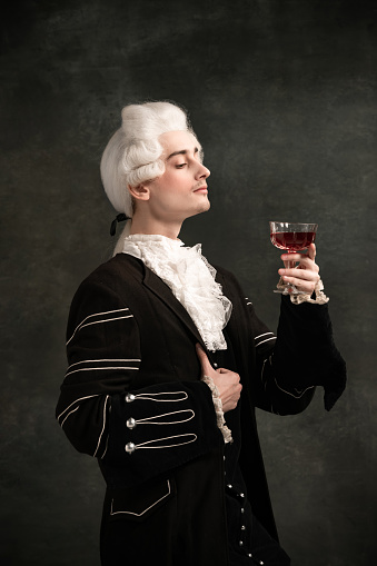 Classic music and wine. Young man wearing wig and vintage medieval outfit like famous composer isolated on dark green vintage background. Retro style, fashion, art, comparison of eras concept.