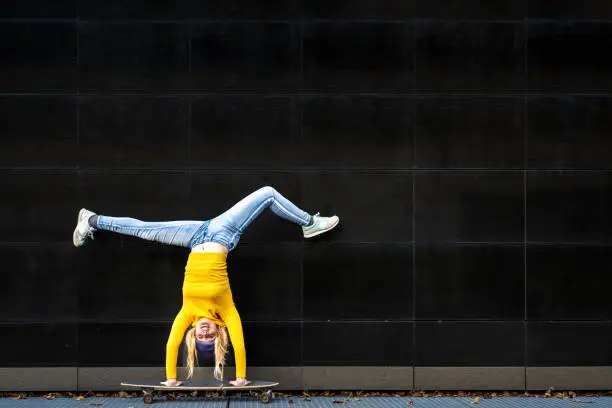 Athelic young woman upside down on a skaboard, black wall background copy space, yellow shirt and blue jeans, generation z female crazy skater lifestyle