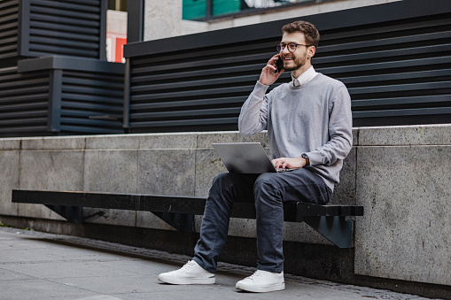 A young man is using a laptop and communicating on the phone while sitting on the bench
