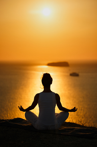 Silhouette of a relaxed woman meditating in Lotus position on a hill above the sea at sunset. Copy space.