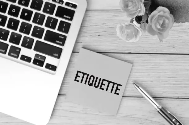 Photo of Black and white picture of laptop, pen, flowers and memo note written with ETIQUETTE. Business and education concept