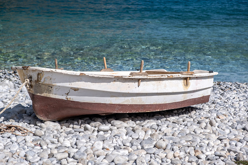 Fishing boat empty, wooden and rusty abandoned on pebble coast. Old vessel moored on shore, calm transparent sea background. Summer destination Greece.