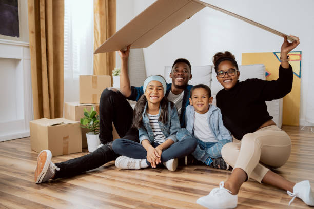 family complete parents with two children sitting on floor relaxing after moving to new apartment holding piece of cardboard left over from boxes as roof over their heads smiling to camera each other - family large american culture fun imagens e fotografias de stock