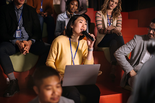 Young woman asking a question while attending business conference in a room full of audience