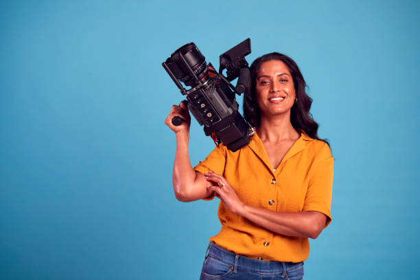 портрет зрелой женщины-видеооператора с камерой, снимающей видео на синем фоне в студии - camera operator стоковые фото и изображения
