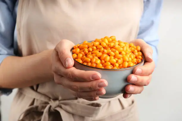 Woman holding bowl with fresh ripe sea buckthorn on light background, closeup