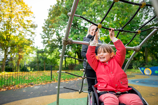 A cheerful low angle medium close-up of a young girl who is a wheelchair user playing in the park on the climbing frame with her mother. They're in a playground ina. public park in Newcastle upon Tyne in the North East of England