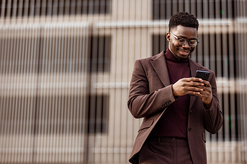 A smiling African businessman in a suit reads a message on his mobile phone while walking around the city
