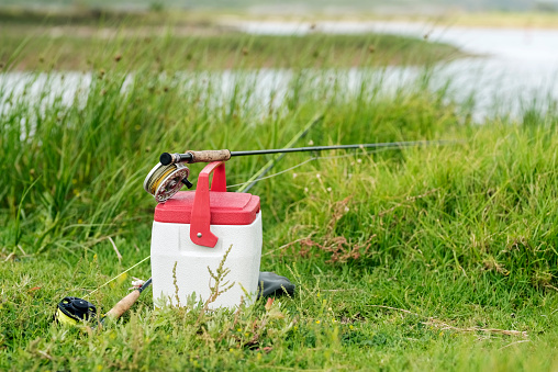 Shot of a fly fishing rod over red and white fishing tackle box on grass along the river bank