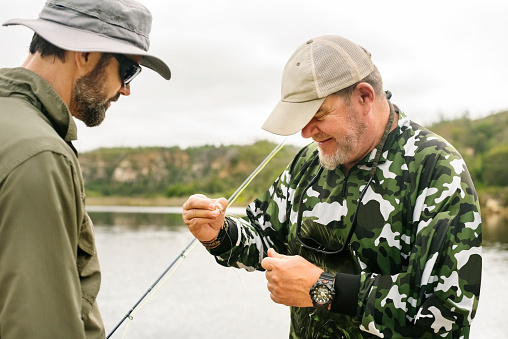 Two mature man wearing hat putting lure on fishing line while standing by the lake