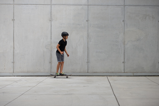 Helmet-fitted brunette teenager skateboard trains on platform in front of concrete building. .  Copy Space.
