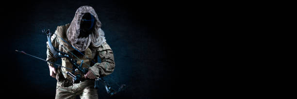 Unrecognizable silent killer with a compound bow in military uniform against a dark concrete wall. Concept of modern military special operations stock photo