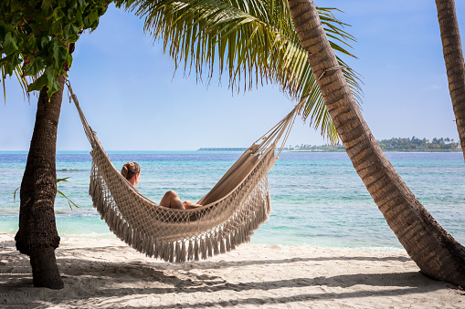 Beach vacation holidays with woman relaxing in hammock between coconut palm tree, white sand, blue sky and turquoise water. Scenic island resort in Maldives, perfect travel destination.