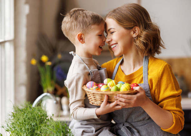 Sweet family portrait of young mother and little son with wicker basket full of painted Easter eggs Sweet family portrait of happy mother and little son holding wicker basket full of painted multi-colored Easter eggs, tenderly embracing and smiling in cozy light kitchen at home, selective focus easter sunday photos stock pictures, royalty-free photos & images