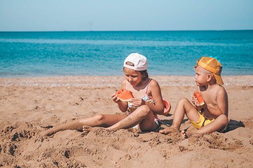 Portrait of children of sister and brother on the beach with watermelon. Girl and little boy are eating on the beach. The children had a picnic by the sea. Copy space.