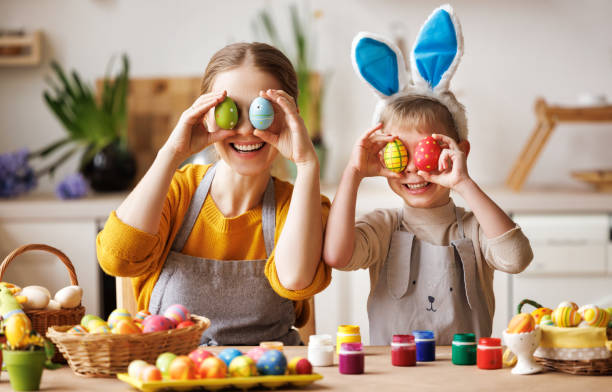 Happy mother and little son sitting at table in kitchen and covering eyes with Easter eggs Happy mother and little son wearing aprons holding painted colorful Easter eggs in front of eyes while decorating them with food dyes in cozy kitchen at home. Easter craft activities for families easter egg coloring stock pictures, royalty-free photos & images