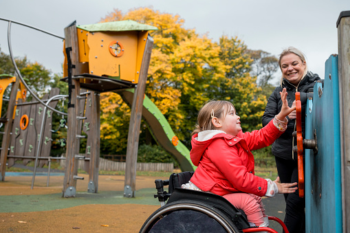 A low angle side view of a mother and her young daughter who is a wheelchair user playing in an accessible playground in a public park in Newcastle upon Tyne in the North East of England,