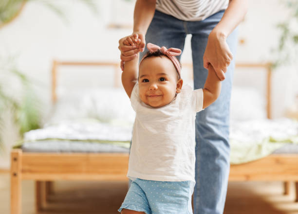 happy ethnic baby girl takes her first steps smiling and holding her mother's hands happy African American baby girl takes her first steps smiling and holding her mother's hands first steps stock pictures, royalty-free photos & images