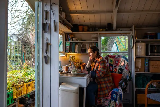 Side view of a woman sitting in her garden summer house cabin using her laptop in Hexham, North East of England.