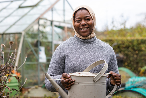 Woman volunteer working outdoors on a community farm, carrying a watering can ready to tend to her crops. The farm is sustainable and environmentally friendly in the North East of England. She is smiling, looking at the camera.
