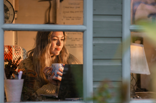 Through window shot of a woman sitting in her garden summer house cabin at night using her laptop in Hexham, North East of England. The is holding a mug with a hot drink.