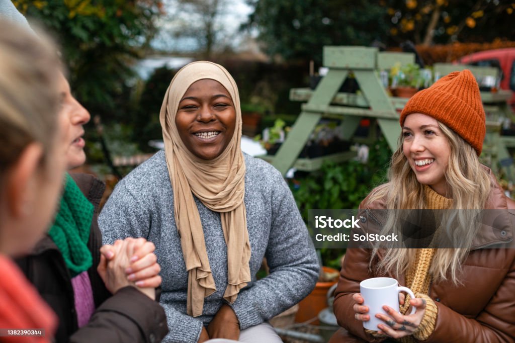 Diverse Women Volunteers Volunteers sitting outdoors wearing warm casual clothing on a sunny cold winters day. They are resting and having a tea break from working on a community farm, looking after crops and performing other sustainable and environmentally friendly tasks. They are laughing and talking together, drinking hot drinks. Community Stock Photo