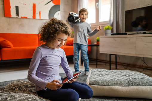 Children having fun at home alone two siblings brother and sister boy holding wifi speaker listening to the music while his sister is playing video games using digital tablet having fun at home