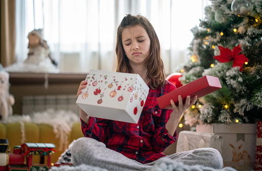 Unhappy girl opening Christmas present while sitting in front of Christmas tree at home