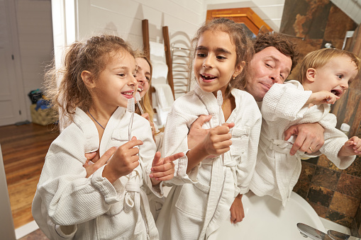 Young happy parents in white bathrobe standing in the bathroom and getting ready to brush their teeth with children