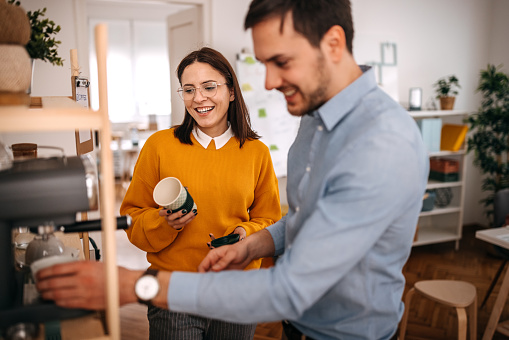 Smiling male and female colleagues making coffee while standing in cafeteria at small office