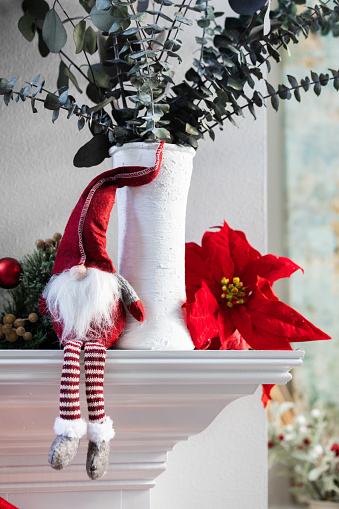 A classic norweigan holiday Christmas gnome decoration sitting on a white modern mantle with a white vase with eucalyptus and a poinsettia bloom.
