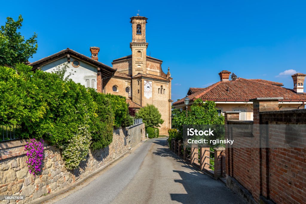 Narrow street and old church in Guarene, Italy. Narrow street, houses with green bushes and old brick church under blue sky in small town of Guarene in Piedmont, Northern Italy. Architecture Stock Photo