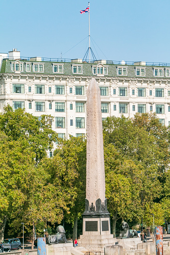 Cleopatra's Needle and Savoy Hotel at Victoria Embankment in City of Westminster, London, with people visible on the riverbank. The obelisk was removed from Egypt by Isma'il Pasha. It was constructed in Heliopolis (Cairo) under the reign of 18th Dynasty Pharaoh Thutmose III.