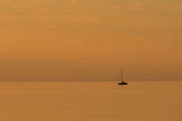 A lonely sailboat is the focus of a minimalist scene during a gold hued sunset on Lake Huron in the summer