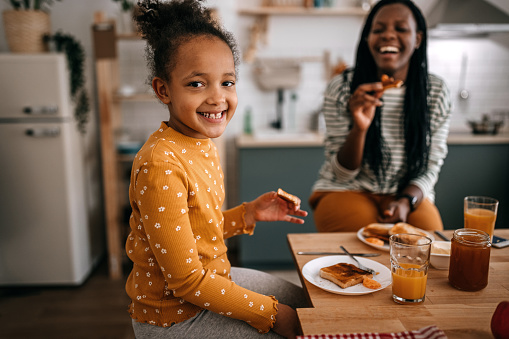 Portrait of smiling cute girl eating breakfast with cheerful mother while sitting at dining table