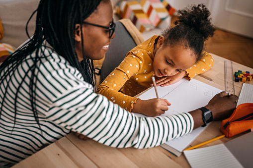 Mid adult mother with dreadlocks assisting daughter in writing homework on table at home
